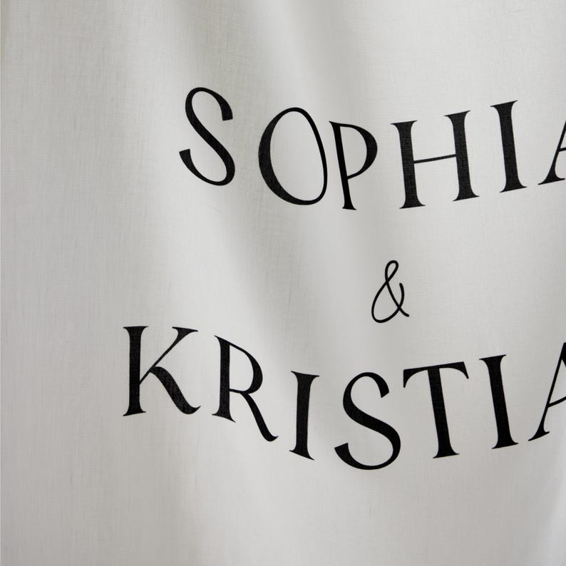 May include: A white fabric banner featuring the black printed text SOPHIA & KRISTIAN. The material has a visible woven texture and soft folds, with the names arranged in two lines separated by an ampersand in a classic serif typeface.