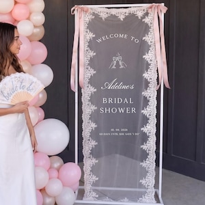 Op de afbeelding: Een rechthoekig wit metalen frame houdt een transparant stoffen paneel vast met witte kanten randen en roze linten. Het paneel bevat witte tekst met WELCOME TO, Adeline's BRIDAL SHOWER, 10. 08. 2026, 60 DAYS UNTIL SHE SAYS I DO en een wijnglasicoon.