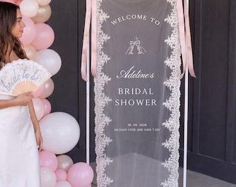 May include: A rectangular white metal frame holds a sheer fabric panel with white lace trim and pink ribbon ties. The panel features white text reading WELCOME TO, Adeline's BRIDAL SHOWER, 10. 08. 2026, 60 DAYS UNTIL SHE SAYS I DO, and a wine glass icon.