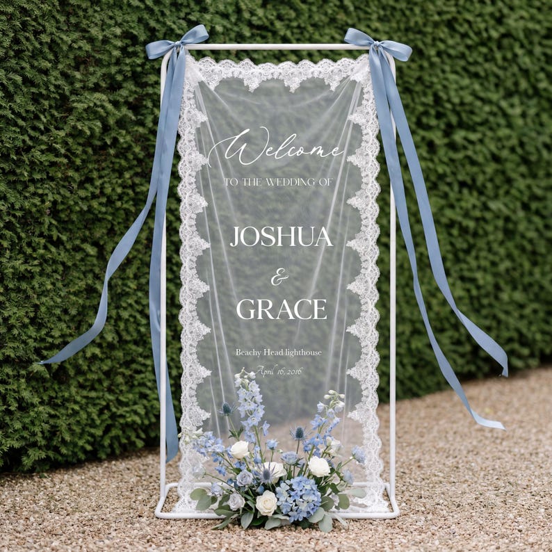 May include: A white metal wedding welcome sign with a sheer, lacy fabric panel. The sign features the names "Joshua & Grace" and is adorned with blue ribbons and a floral arrangement of blue and white flowers.