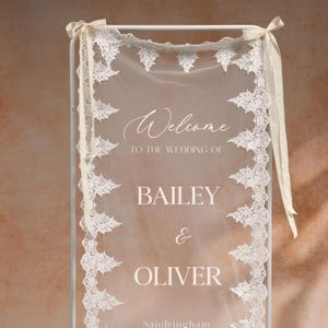 May include: A wedding welcome sign with the names Bailey & Oliver. The sign features a sheer white fabric with delicate lace trim and the word "Welcome" in elegant script. Cream-colored ribbons adorn the top.