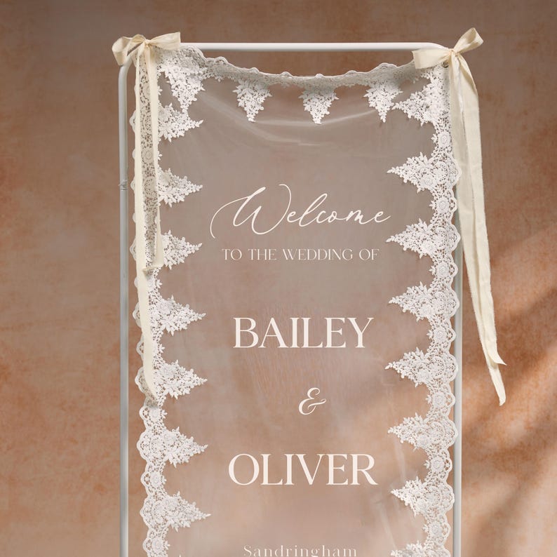 May include: A wedding welcome sign with the names Bailey & Oliver. The sign features a sheer white fabric with delicate lace trim and the word "Welcome" in elegant script. Cream-colored ribbons adorn the top.