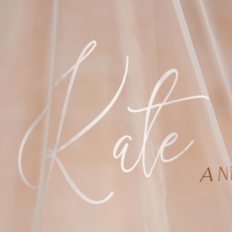 May include: A sheer, light-colored fabric with the name "Kate" in elegant white script. The fabric has a soft, translucent quality, with the word "AND" visible in the background.