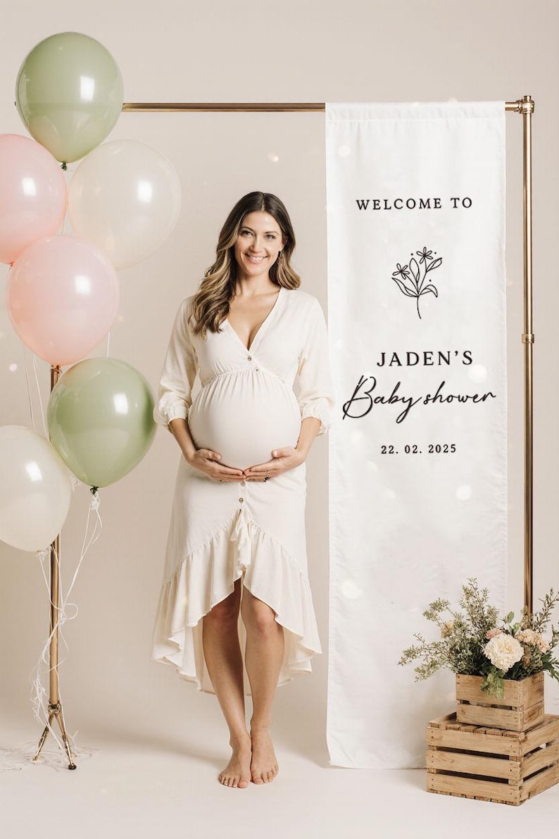 May include: A pregnant person in a white dress stands in front of a banner that reads "Welcome to Jaden's Babyshower 22.02.2025." Balloons in shades of green, pink, and white are tied to a stand. Wooden crates hold flowers.