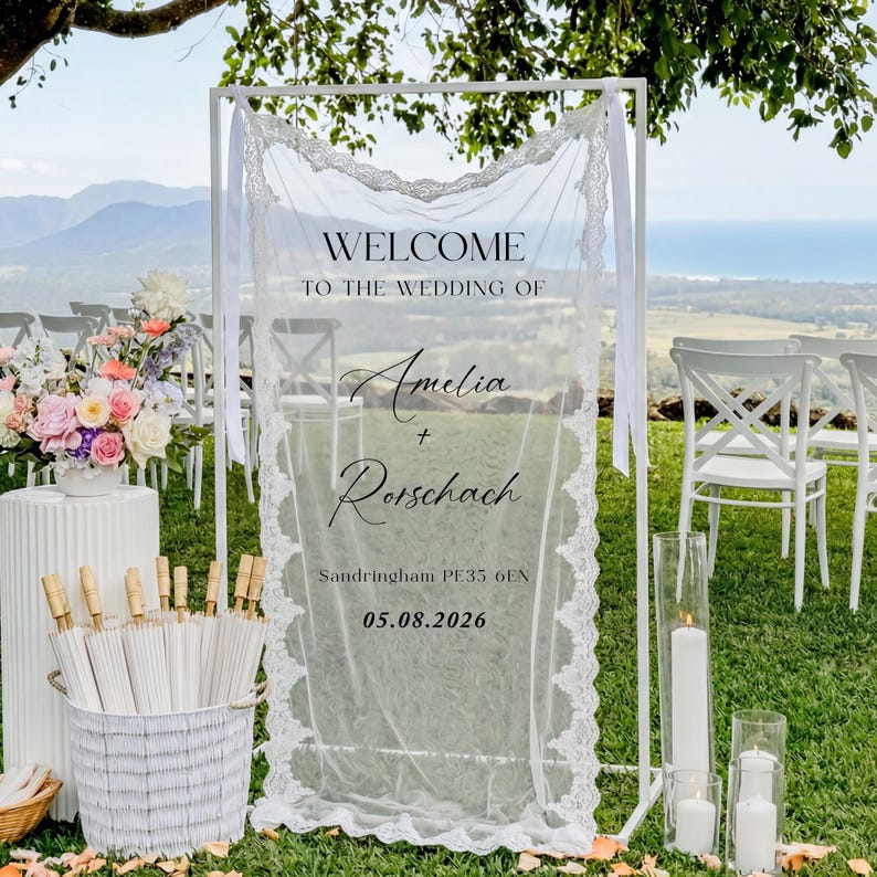 May include: A white wedding welcome sign with the text "Welcome to the Wedding of Amelia + Rorschach" and the date 05.08.2026. The sign is decorated with lace and ribbons, set against a scenic outdoor backdrop.