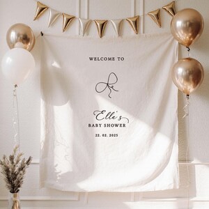 May include: A baby shower backdrop with a white banner featuring the text "Welcome to Elle's Baby Shower 22.02.2025". The banner is decorated with gold balloons and a gold triangle banner.