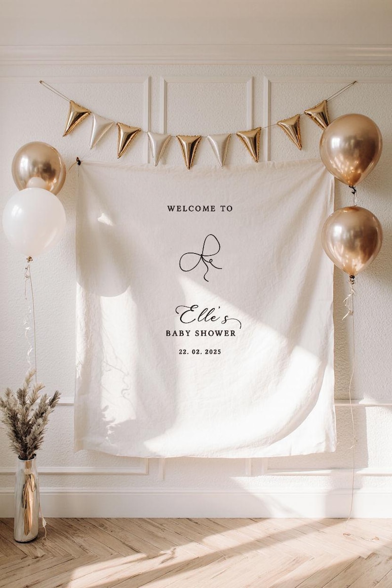 May include: A baby shower backdrop with a white banner featuring the text "Welcome to Elle's Baby Shower 22.02.2025". The banner is decorated with gold balloons and a gold triangle banner.