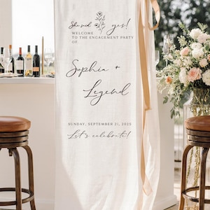May include: A vertical off-white fabric banner hanging from a rod with a beige ribbon tied at the top right. The banner features black text reading "She said yes! WELCOME TO THE ENGAGEMENT PARTY OF Sophia + Legend SUNDAY, SEPTEMBER 21, 2025 Let's celebrate!"