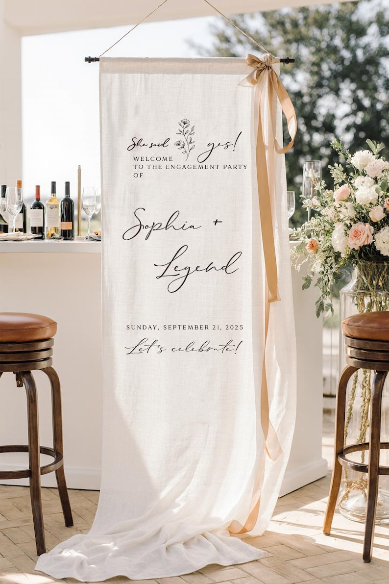 May include: A vertical off-white fabric banner hanging from a rod with a beige ribbon tied at the top right. The banner features black text reading "She said yes! WELCOME TO THE ENGAGEMENT PARTY OF Sophia + Legend SUNDAY, SEPTEMBER 21, 2025 Let's celebrate!"