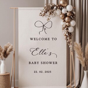 May include: A baby shower welcome sign with a bow graphic, the text "Welcome to Elle's Baby Shower 22.02.2025", and a decorative balloon arch in gold and white. The sign is on a wooden stand.