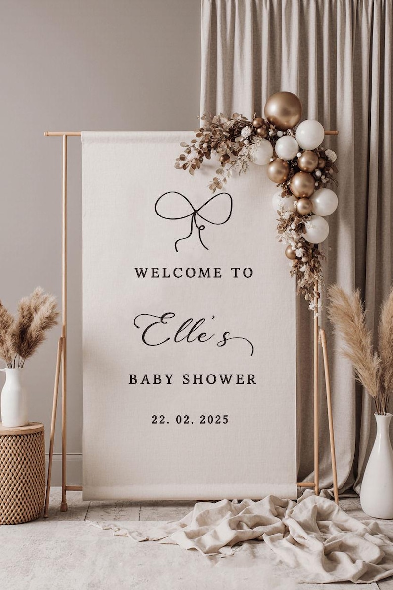May include: A baby shower welcome sign with a bow graphic, the text "Welcome to Elle's Baby Shower 22.02.2025", and a decorative balloon arch in gold and white. The sign is on a wooden stand.