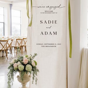 May include: A tall, off-white fabric banner hanging from a metal frame, featuring the text "we're engaged", "WELCOME TO THE ENGAGEMENT PARTY OF", "SADIE and ADAM", "SUNDAY, SEPTEMBER 21, 2025", and "THE BROADMOOR". Green ribbons are tied at the top corners.