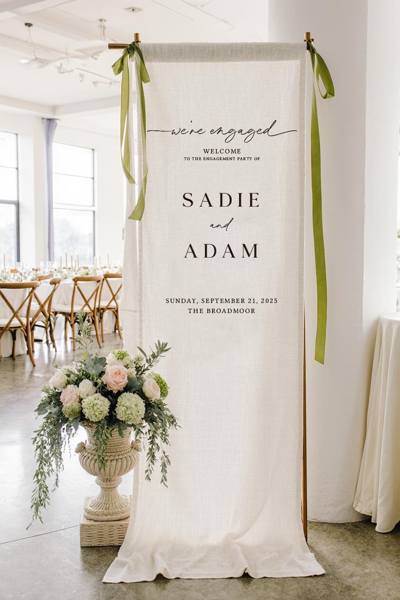 May include: A tall, off-white fabric banner hanging from a metal frame, featuring the text "we're engaged", "WELCOME TO THE ENGAGEMENT PARTY OF", "SADIE and ADAM", "SUNDAY, SEPTEMBER 21, 2025", and "THE BROADMOOR". Green ribbons are tied at the top corners.