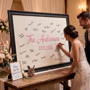 May include: A large rectangular guest book sign in a black frame featuring a cream-colored fabric background with the pink embroidered text The Andersons and 07.01.2026, surrounded by numerous handwritten signatures in black ink on the fabric.