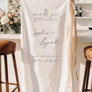 May include: A vertical white fabric banner hanging from a wooden rod with beige ribbon accents. The text reads: She said yes! WELCOME TO THE ENGAGEMENT PARTY OF Sophia + Legend SUNDAY, SEPTEMBER 21, 2025 Let's celebrate! It features a small floral line drawing.