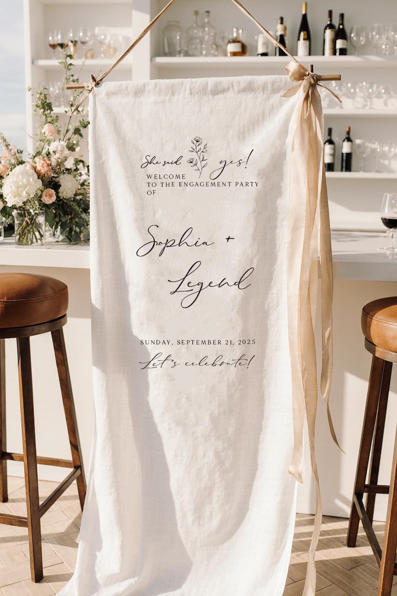 May include: A vertical white fabric banner hanging from a wooden rod with beige ribbon accents. The text reads: She said yes! WELCOME TO THE ENGAGEMENT PARTY OF Sophia + Legend SUNDAY, SEPTEMBER 21, 2025 Let's celebrate! It features a small floral line drawing.