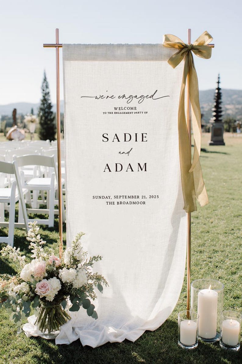 May include: A vertical off-white fabric banner suspended from a copper-colored metal frame. The banner features black text reading "we're engaged", "WELCOME TO THE ENGAGEMENT PARTY OF", "SADIE and ADAM", and "SUNDAY, SEPTEMBER 21, 2025 THE BROADMOOR". A gold ribbon is tied to the top right corner.
