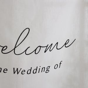 May include: A white fabric sign featuring black text. The word Welcome is written in a large, flowing cursive script, while the phrase the Wedding of is printed below in a smaller, clean, sans-serif font. The material has a visible weave texture.