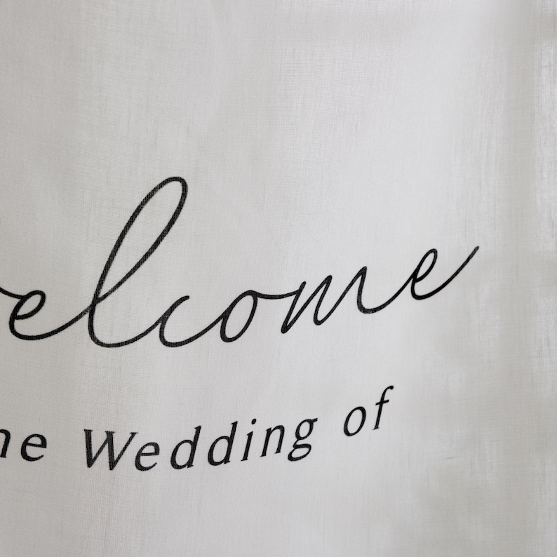 May include: A white fabric sign featuring black text. The word Welcome is written in a large, flowing cursive script, while the phrase the Wedding of is printed below in a smaller, clean, sans-serif font. The material has a visible weave texture.