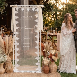 May include: A wedding welcome sign with the names "Amelia + James" and the date 05.08.2026. The sign is framed in white and adorned with lace and ribbons. The scene includes floral arrangements and a couple.