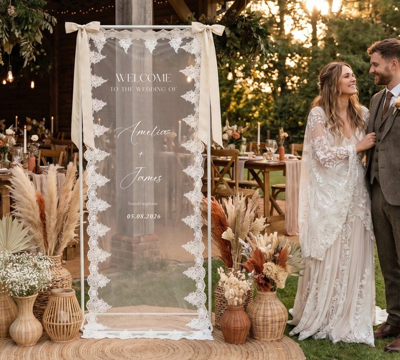 May include: A wedding welcome sign with the names "Amelia + James" and the date 05.08.2026. The sign is framed in white and adorned with lace and ribbons. The scene includes floral arrangements and a couple.