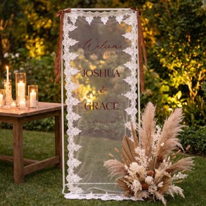 May include: A wedding welcome sign with the names Joshua & Grace, framed in white with a lace border. The sign is transparent, with brown text and ribbons. A floral arrangement and candles are in the background.