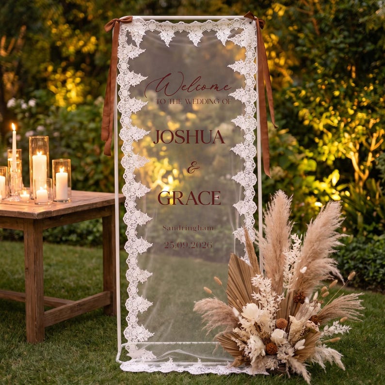 May include: A wedding welcome sign with the names Joshua & Grace, framed in white with a lace border. The sign is transparent, with brown text and ribbons. A floral arrangement and candles are in the background.