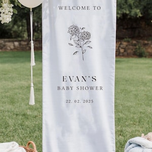 May include: White fabric banner with the text "WELCOME TO EVAN'S BABY SHOWER 22.02.2025" in black font, along with a floral illustration. The banner is hanging outdoors, with white balloons and greenery in the background.