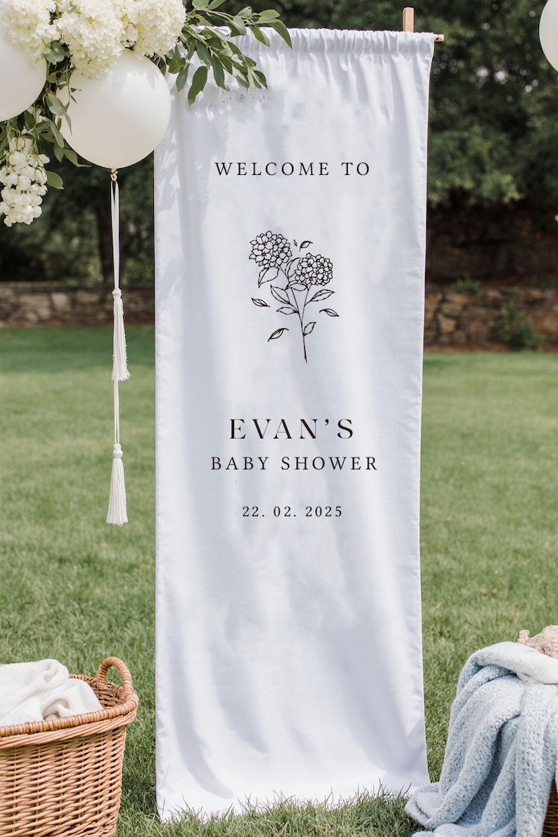 May include: White fabric banner with the text "WELCOME TO EVAN'S BABY SHOWER 22.02.2025" in black font, along with a floral illustration. The banner is hanging outdoors, with white balloons and greenery in the background.