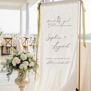 May include: A tall, off-white fabric banner hanging from a copper-colored metal frame, featuring black printed text that reads: She said yes! Welcome to the engagement party of Sophia + Legend, Sunday, September 21, 2025, Let's celebrate!