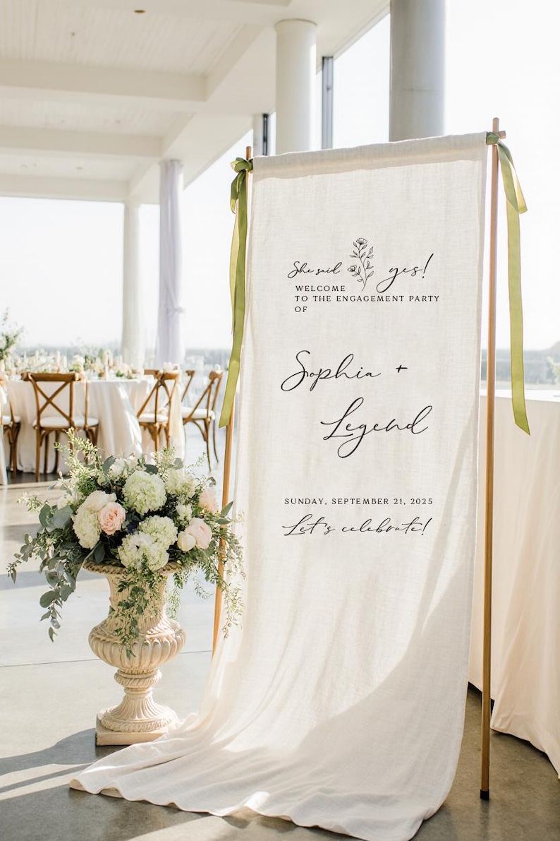 May include: A tall, off-white fabric banner hanging from a copper-colored metal frame, featuring black printed text that reads: She said yes! Welcome to the engagement party of Sophia + Legend, Sunday, September 21, 2025, Let's celebrate!