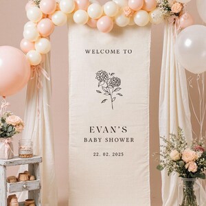 May include: A baby shower backdrop featuring a cream-colored banner with the text "WELCOME TO EVAN'S BABY SHOWER 22. 02. 2025". The banner is decorated with floral arrangements, balloons in white and pink, and draped fabric.