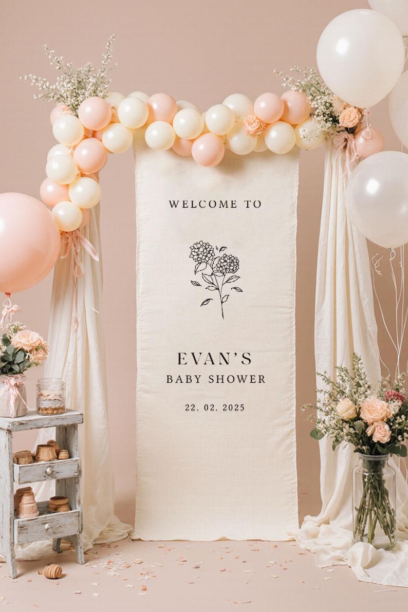 May include: A baby shower backdrop featuring a cream-colored banner with the text "WELCOME TO EVAN'S BABY SHOWER 22. 02. 2025". The banner is decorated with floral arrangements, balloons in white and pink, and draped fabric.