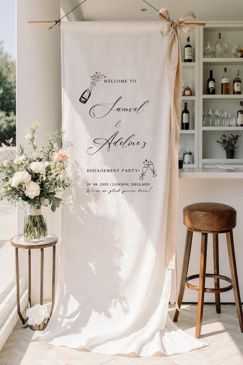 May include: A tall, off-white fabric banner hanging from a gold rod with a beige ribbon tie. The banner features black text reading: WELCOME TO Samuel & Adeline's ENGAGEMENT PARTY! 10. 08. 2025 | LONDON, ENGLAND We're so glad you're here! with illustrations.