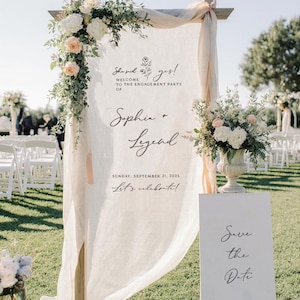 May include: A tall wooden frame draped with sheer cream fabric, featuring floral arrangements and text reading: She said yes! WELCOME TO THE ENGAGEMENT PARTY OF Sophia + Legend SUNDAY, SEPTEMBER 21, 2025 Let's celebrate! Beside it stands a white sign: Save the Date.