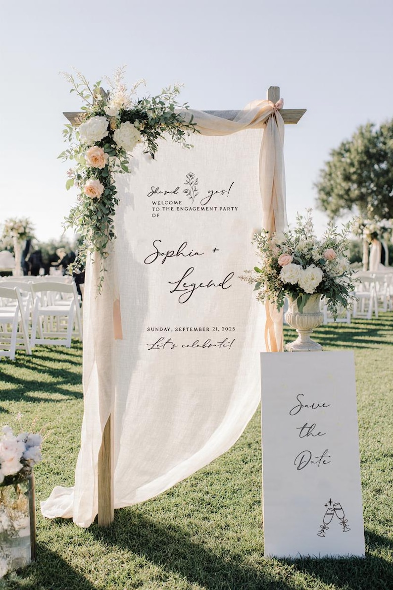 May include: A tall wooden frame draped with sheer cream fabric, featuring floral arrangements and text reading: She said yes! WELCOME TO THE ENGAGEMENT PARTY OF Sophia + Legend SUNDAY, SEPTEMBER 21, 2025 Let's celebrate! Beside it stands a white sign: Save the Date.