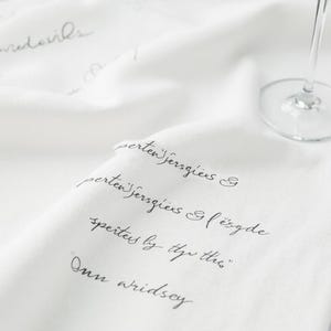 May include: White linen tablecloth with elegant script writing in gray. The fabric is draped, with a wine glass visible in the background. The script appears to be a quote or poem.