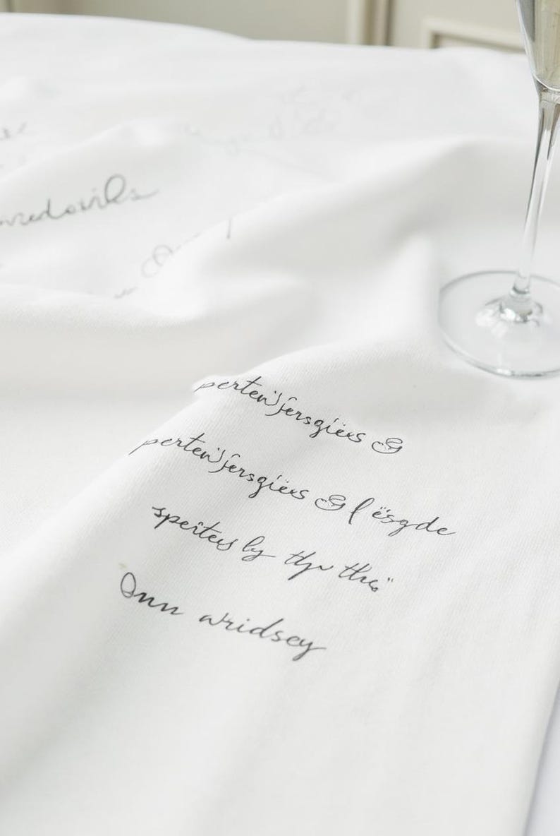 May include: White linen tablecloth with elegant script writing in gray. The fabric is draped, with a wine glass visible in the background. The script appears to be a quote or poem.
