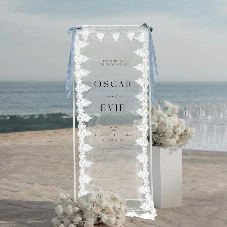 May include: A clear acrylic wedding welcome sign with white lace trim and light blue ribbons. The sign reads "Welcome to the Wedding of Oscar and Evie" with the wedding date and location at the bottom. White flowers are in the foreground.