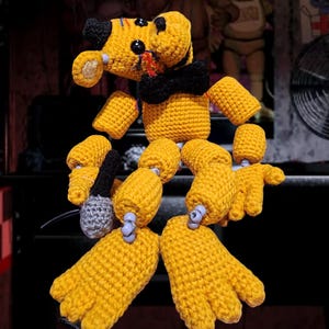 May include: Two crocheted Golden Freddy figures from FNAF, in a marionette style with open mouths. The figures are yellow with black accents and articulated limbs. The image includes text that reads "Golden Freddy from FNAF" and "Updated pattern January 6th, 2026."