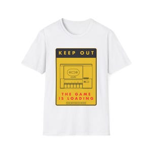 Może przedstawiać: Biały t-shirt z krótkim rękawem z prostokątną grafiką na żółtym tle z czarną obwódką. Grafika przedstawia rysunek liniowy odtwarzacza kaset, czarny napis KEEP OUT, czerwony napis THE GAME IS LOADING oraz napis FOR[RETRO]LOOP.