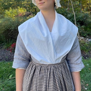 May include: A person wearing a historical costume. The outfit includes a white collar, gray long-sleeve shirt, and a black and white checkered skirt. A white bonnet is also worn. The clothing is made of fabric.