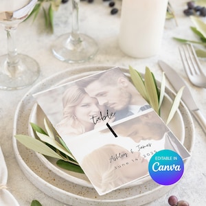 Puede incluir: Una tarjeta con el número de mesa de boda con una foto de una pareja, la palabra "mesa" y el número "1". La tarjeta también incluye los nombres "Ashley & James" y la fecha "14 de junio de 2025". La tarjeta está sobre un plato blanco con vegetación.