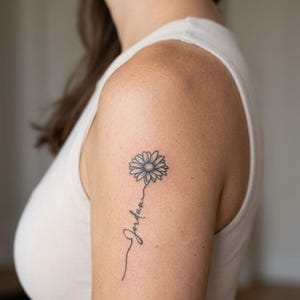 May include: A black ink tattoo on an upper arm featuring a daisy flower with a thin stem that transitions into the cursive script name Jordan. The design is rendered in fine lines with a single flower head and a delicate, flowing stem.