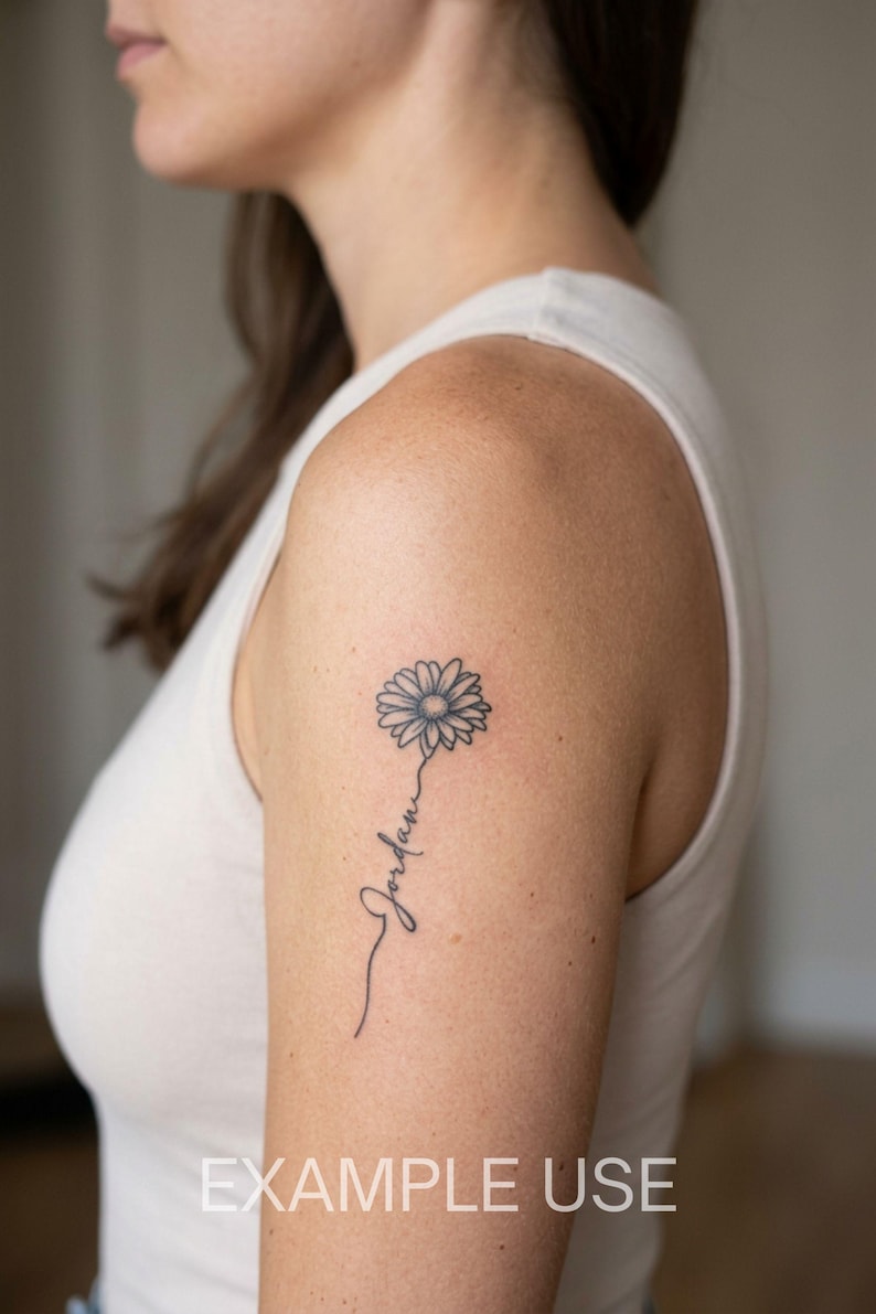 May include: A black ink tattoo on an upper arm featuring a daisy flower with a thin stem that transitions into the cursive script name Jordan. The design is rendered in fine lines with a single flower head and a delicate, flowing stem.