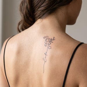 May include: A black ink tattoo on the upper back featuring a delicate lily of the valley flower stem that transitions into a cursive script name. The design is rendered in fine lines, positioned vertically between the shoulder blades.