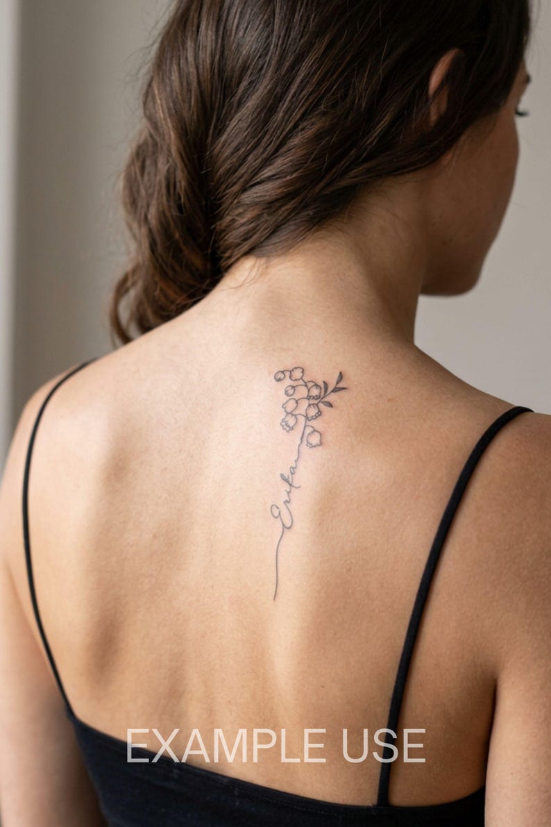 May include: A black ink tattoo on the upper back featuring a delicate lily of the valley flower stem that transitions into a cursive script name. The design is rendered in fine lines, positioned vertically between the shoulder blades.