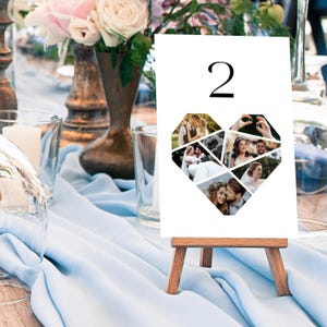 May include: A white table number card with the number 2 in black, featuring a heart-shaped collage of wedding photos. The card is displayed on a small wooden easel, set on a light blue tablecloth.