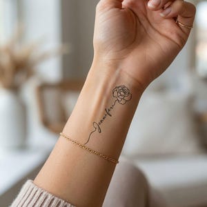 May include: A temporary tattoo on a forearm featuring a black line art drawing of a rose connected to the name Jennifer in a cursive script font. The design is positioned vertically along the inner wrist, accompanied by a thin gold chain bracelet.
