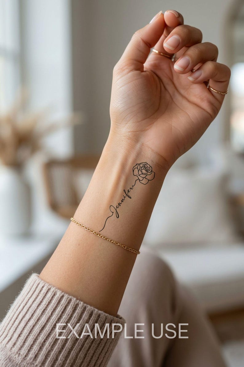 May include: A temporary tattoo on a forearm featuring a black line art drawing of a rose connected to the name Jennifer in a cursive script font. The design is positioned vertically along the inner wrist, accompanied by a thin gold chain bracelet.